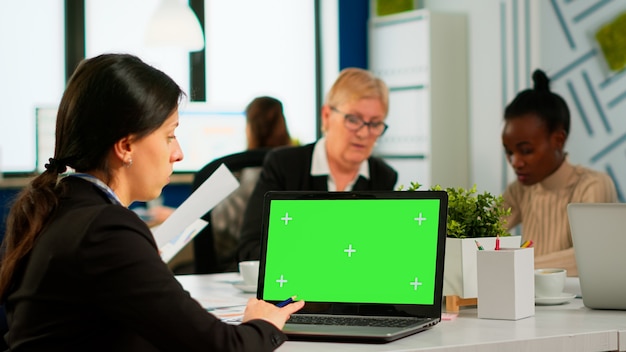 Photo over the sholder of manager sitting at meeting desk looking at laptop with green screen mockup while diverse team working on background. multiethnic people planning project on chroma key display