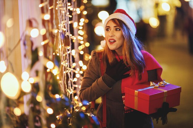 Shoot of a surprised young woman with red present box is having fun in the city street at Christmas time.