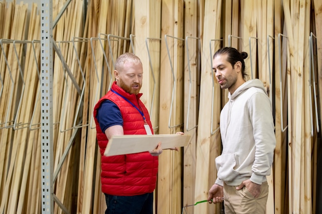Продавец строительного магазина помогает молодому человеку выбрать деревянную доску