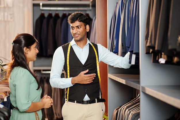 Shop assistant showing young woman assortment of jackets and male suit in store