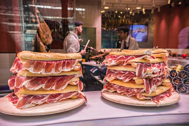 Shop showcase selling jamon spanish ham sandwiches, pile of\
bread with ham snacks for sale, zaragoza