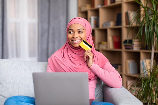 Shopaholic dreamy black muslim lady holding credit card and using laptop thinking about purchases