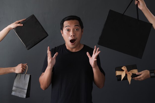 Shopaholic man surrounded by many hands holding shopping bags with surprised expression