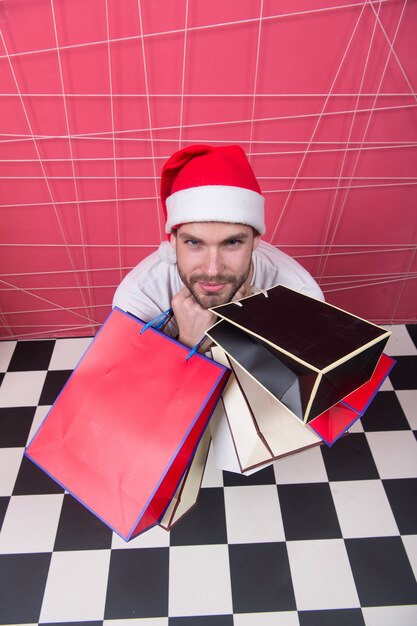 Shopper in red hat with paperbags, top view. Man santa hold bags on black and white checkered floor. Christmas, new year presents. Winter sale, shopping concept. Holidays preparation and celebration.