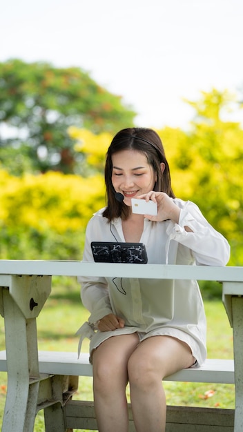 オンラインで購入するか、夏に都市公園のベンチに座っているスマートフォンとクレジットカードでオンラインで作業する買い物客の笑顔の女の子笑顔の女の子は、エグゼクティブの若い彼女のカードの肖像画に満足しています