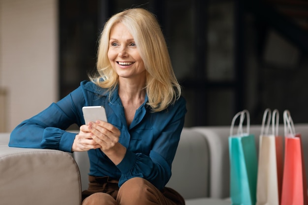 Shopping app happy mature woman using cellphone purchasing new clothes online in app sitting on sofa