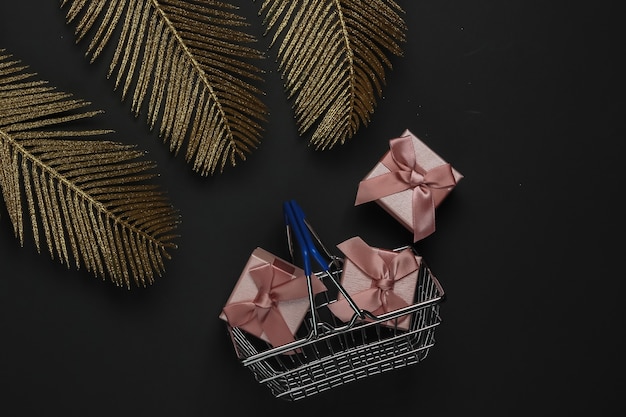 Shopping basket with gift box on black background with golden palm leaves. Fashion flat lay. Black Friday. Top view