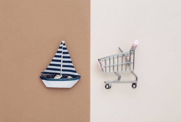 Shopping cart and sailboat on brown beige background. Top view