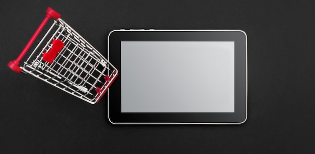 Shopping cart and tablet PC with blank screen on black background Top view Copy space