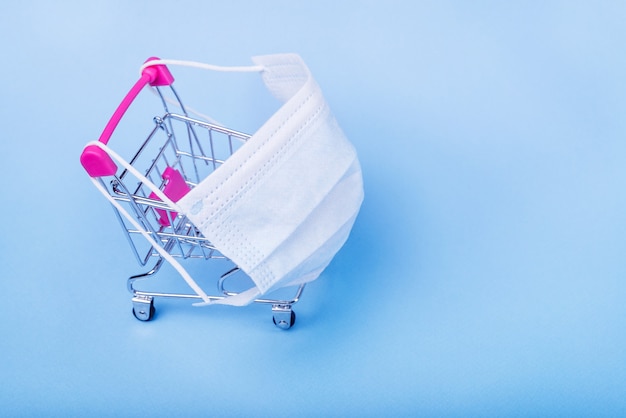 Shopping cart with protective medical mask against coronavirus. Safe and online shopping on quarantine concept. Light blue background, copy space.