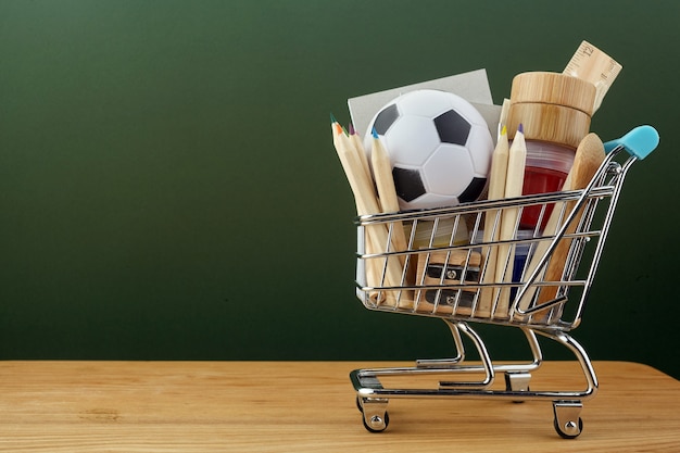 Shopping cart with school supplies over green chalkboard