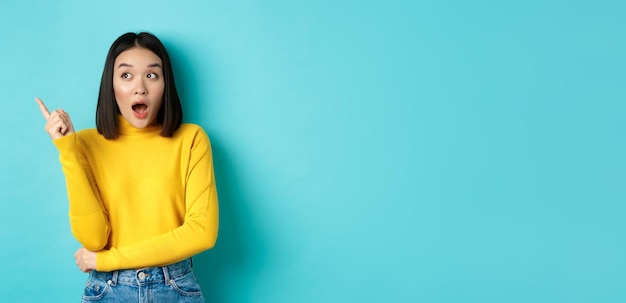 Shopping concept impressed asian girl in yellow pullover pointing and looking left amazed showing lo