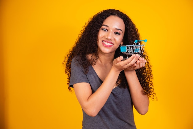 Shopping concept. Portrait of smiling woman holding small empty shopping cart on her palm, isolated on yellow background