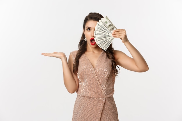 Концепция покупок. Шокированная женщина, держащая деньги возле лица, в стильном платье, показывая доллары с удивленным выражением лица, стоя на белом фоне.