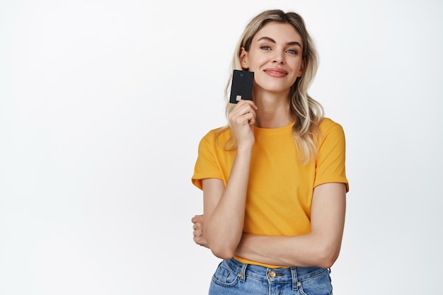 Shopping concept Smiling young woman thinking of buying something holding credit card near face standing over white background