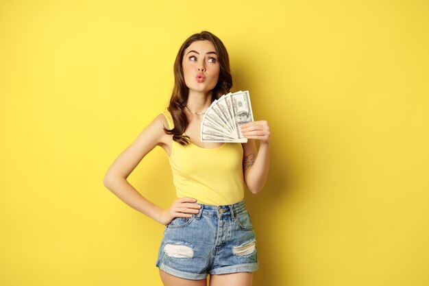 ショッピング、クレジット、お金の概念。若いブルネットの女性は、黄色の背景の上に立って、現金を示し、満足して笑っています。