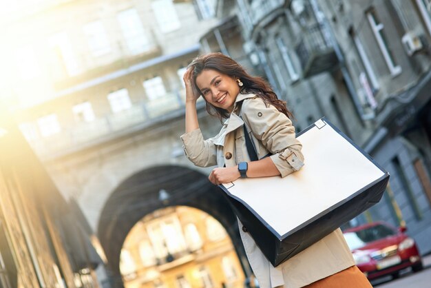 買い物の日。街の通りに立って、カメラを見て、笑顔の大きな買い物袋を持つ若い魅力的でスタイリッシュな女性。ファッション、人々のライフスタイルのコンセプト