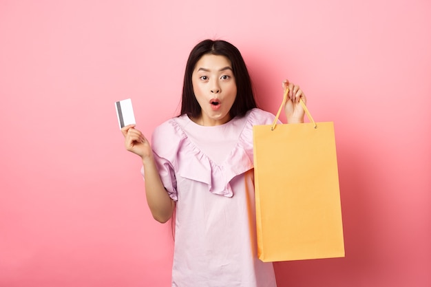 Shopping. Excited asian woman gasping amazed, holding bag from shop and plastic credit card, standing on pink background.