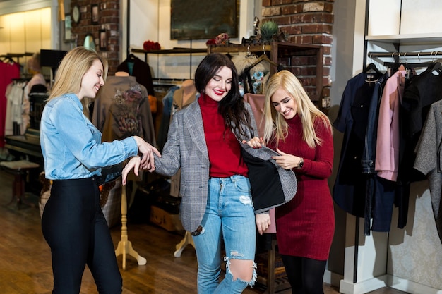 Shopping, fashion and friendship-three smiling friends try on clothes, a jacket in a shopping center.