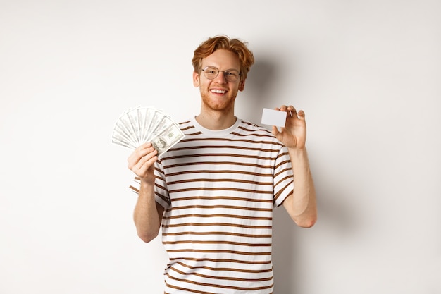 Концепция покупок и финансов. Молодой рыжий мужчина с бородой и очками показывает пластиковую кредитную карту с деньгами в долларах, белый фон