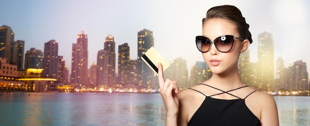 shopping, finances, fashion, people and luxury concept - beautiful young woman in elegant black sunglasses with credit card over dubai night city lights street background