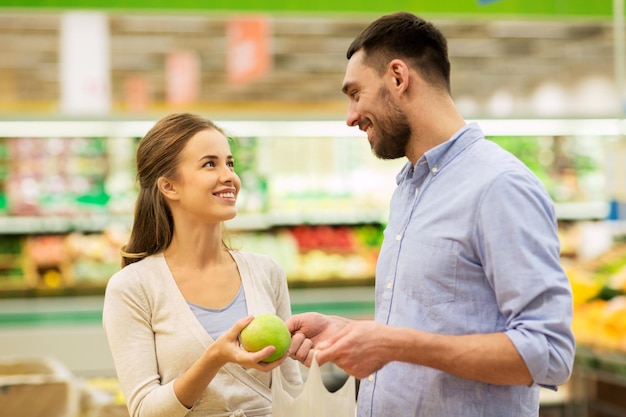 покупки, еда, продажа, потребление и концепция людей - счастливая пара покупает яблоки в продуктовом магазине или супермаркете