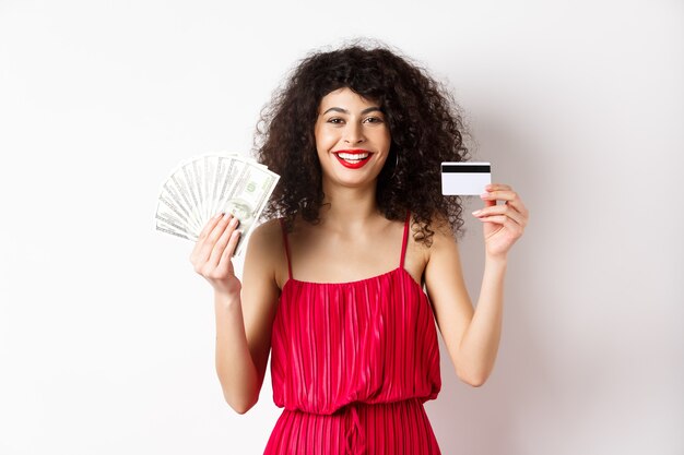 Shopping. Happy beautiful woman with plastic credit card and dollar gills, smiling and looking at camera, wearing red dress, white background.
