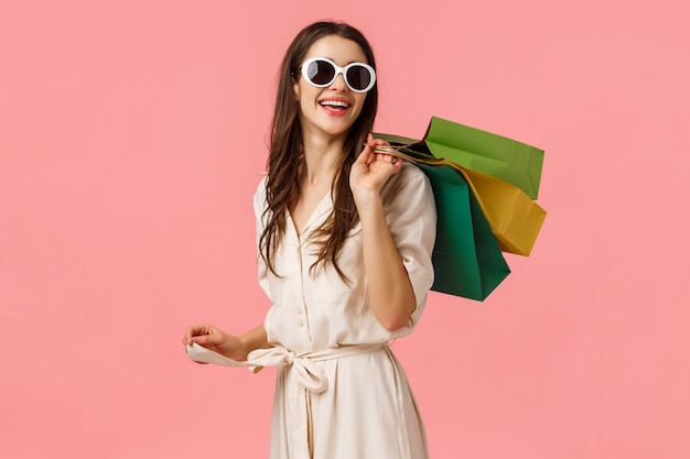 Shopping, lifestyle and consumer concept. Attractive young carefree girl in glasses and dress, look behind with satisfied smile after good shopping, holding bags on back and standing pink wall