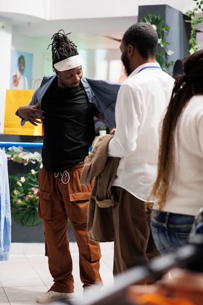 Shopping mall assistant helping customer to try clothes before buying in fashion showroom. Young african american man wearing shirt while checking apparel size in modern store