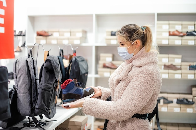 Shopping mall, shoe department. New normal. blonde woman in fake fur with dyed hair in outer clothing, protection medical mask holding black shoe for man