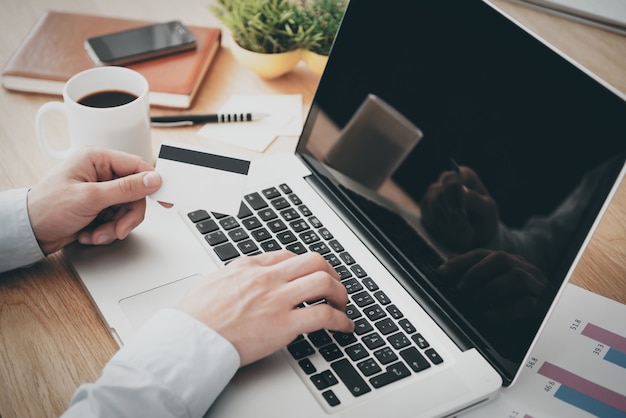 Shopping online. Close-up top view of man working on laptop and holding credit card while sitting at the wooden desk