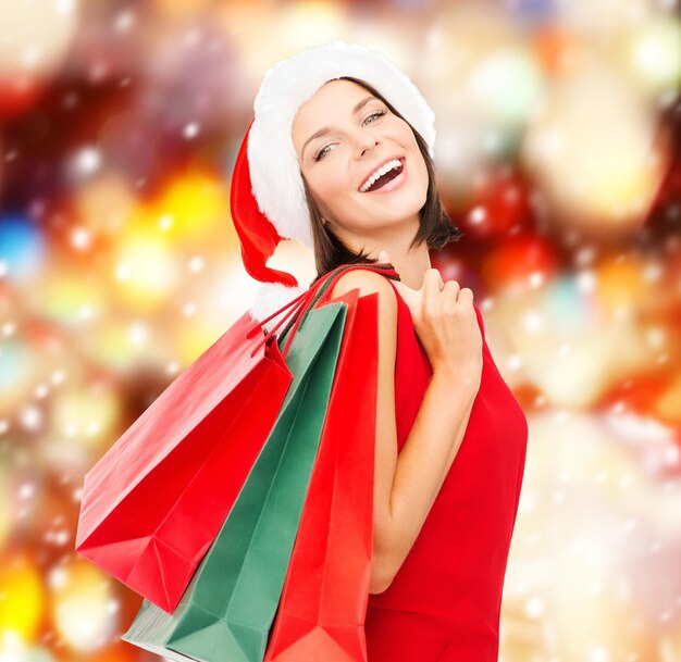 shopping, sale, gifts, christmas concept - smiling woman in red dress and santa helper hat with shopping bags