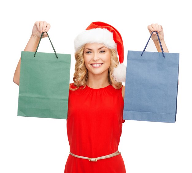 покупки, распродажа, подарки, рождество, рождественская концепция - улыбающаяся женщина в красном платье и шляпе санта-помощника с сумками для покупок