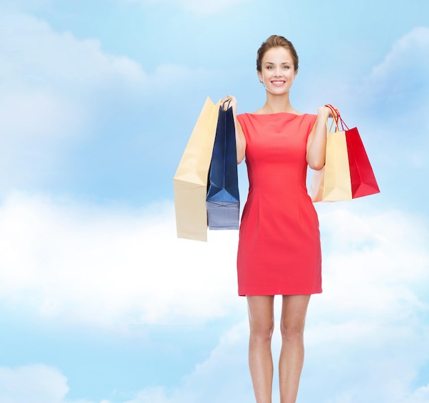 쇼핑, 판매, 휴일 개념 - 파란 흐린 하늘 배경 위에 쇼핑백을 들고 빨간 드레스를 입은 우아한 여성