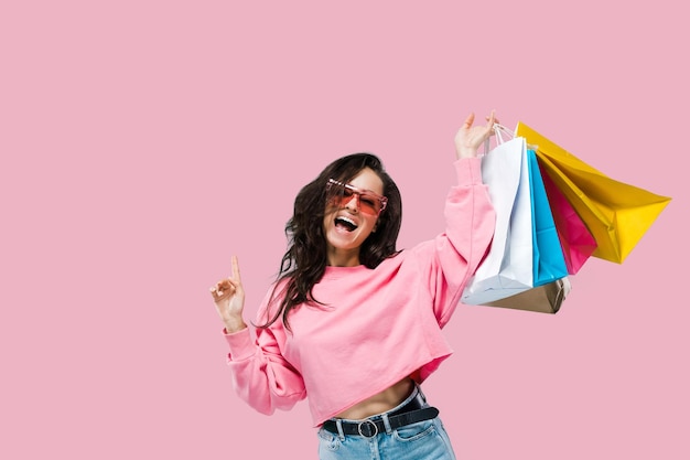 ショッピングと販売のコンセプト カジュアルな服装と孤立したピンクの背景にポーズをとって買い物袋を保持しているピンクのサングラスを着た陽気なトレンディなかなり白人の若い女性は、カメラの笑顔を見ます