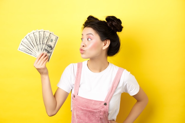 Shopping. Silly korean girl with glam makeup, pucker lips and looking at dollar bills, wasting money, standing on yellow.