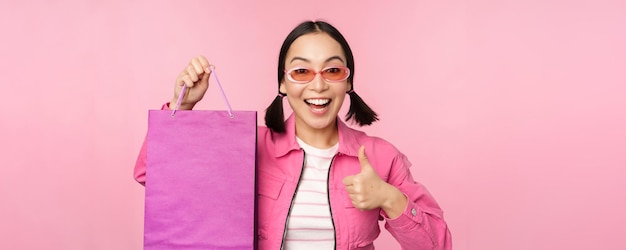 Shopping Stylish asian girl in sunglasses showing bag from shop and smiling recommending sale promo in store standing over pink background