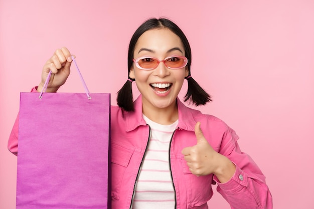 Shopping Stylish asian girl in sunglasses showing bag from shop and smiling recommending sale promo in store standing over pink background