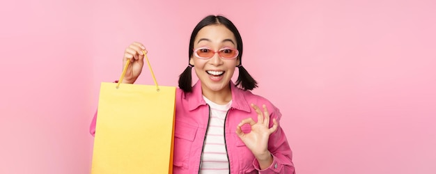 Shopping Stylish asian girl in sunglasses showing bag from shop and smiling recommending sale promo in store standing over pink background