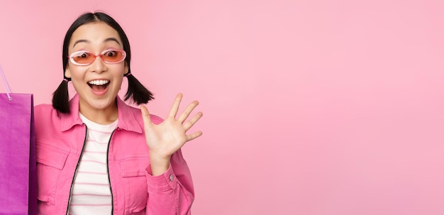 Шопинг Стильная азиатская девушка в солнечных очках показывает сумку из магазина и улыбается, рекомендуя промо-акцию в магазине, стоящую на розовом фоне