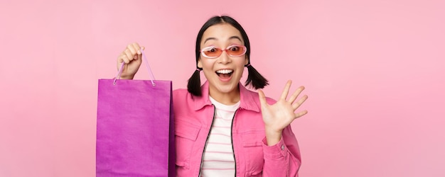 Shopping stylish asian girl in sunglasses showing bag from shop and smiling recommending sale promo