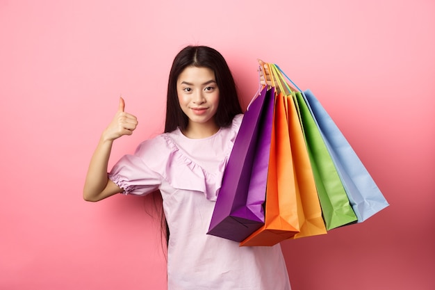 Shopping. ragazza alla moda che mostra i sacchetti di carta colorati e il pollice in su, consiglia sconti del negozio, in piedi su sfondo rosa.