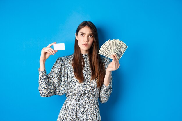 Shopping. thoughtful girl looking up and biting lip, counting in mind or thinking, holding plastic credit card with dollar bills, standing on blue.