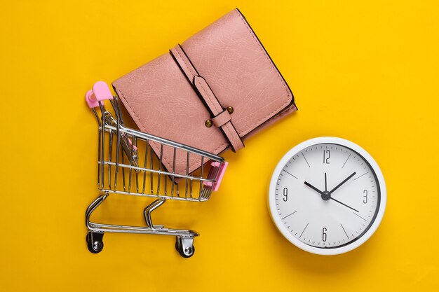 Shopping time. Supermarket trolley with wallet, clock on a yellow background. Minimalism. Top view