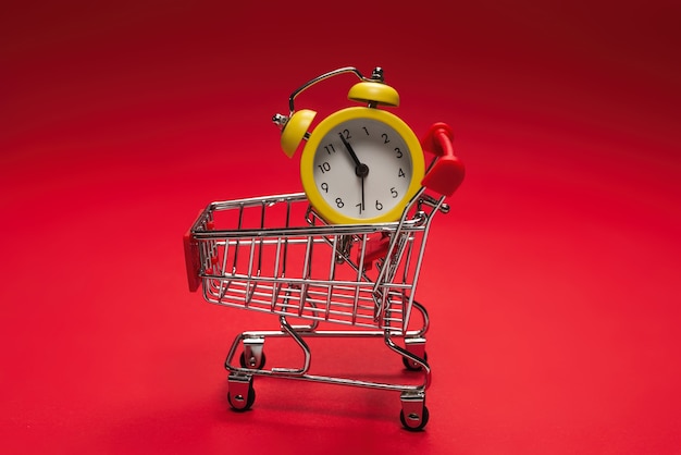 Shopping time. Yellow alarm clock in shopping basket on a red background. Copy space.