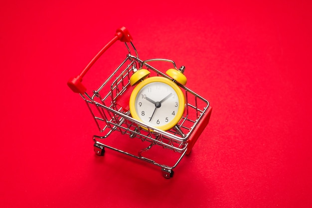 Shopping time. Yellow alarm clock in shopping basket on a red background. Copy space.