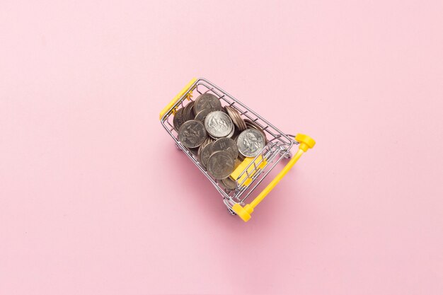 Shopping trolley from a supermarket full of coins on a pink background.