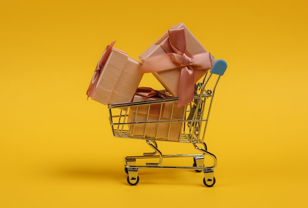 Shopping trolley and gift boxes with bows on yellow background. Composition for christmas, birthday or wedding.