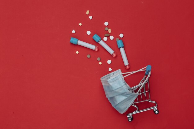 Shopping trolley in medical mask with test tubes, pills on red background. Healthcare