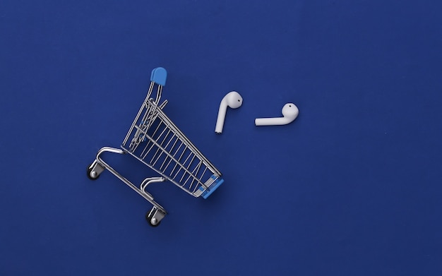 Shopping trolley and modern wireless earphones on classic blue background.